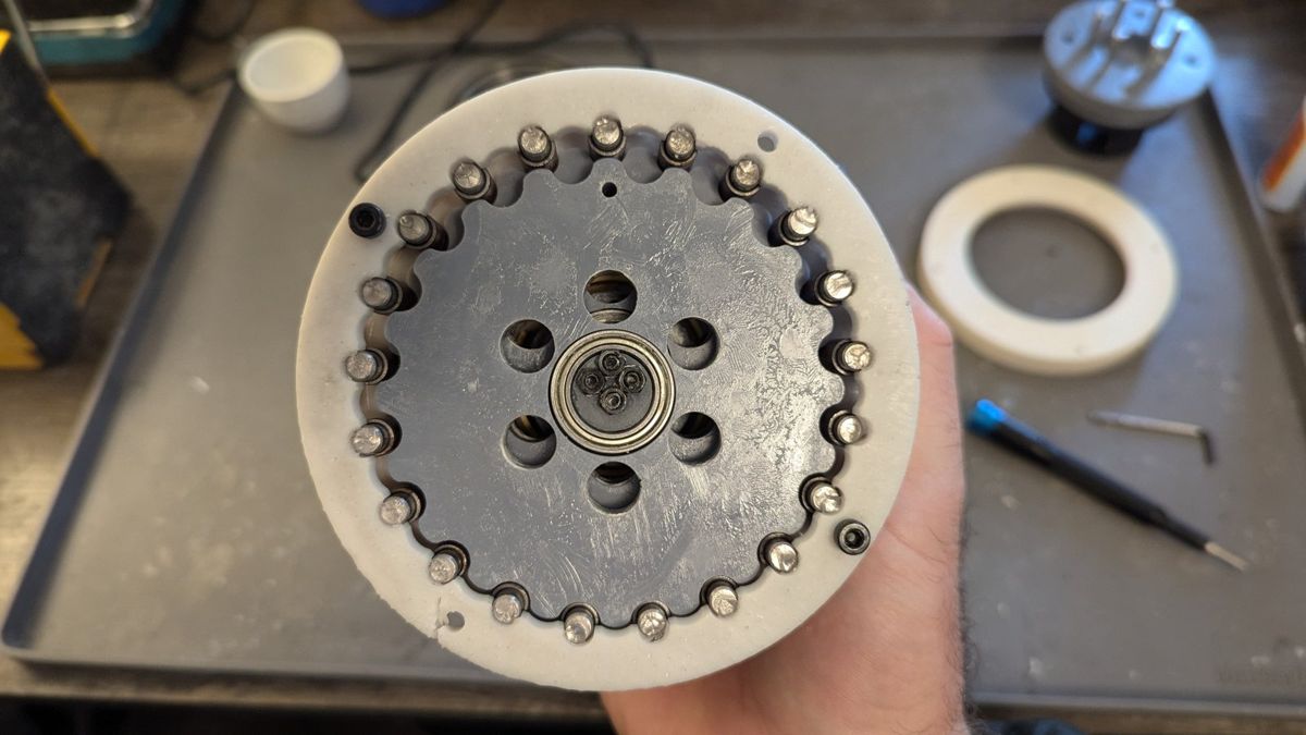 Hand holding the OpenCycloid cycloidal stage: a printed disk running on a ring of 6 mm pins around an eccentric bearing.